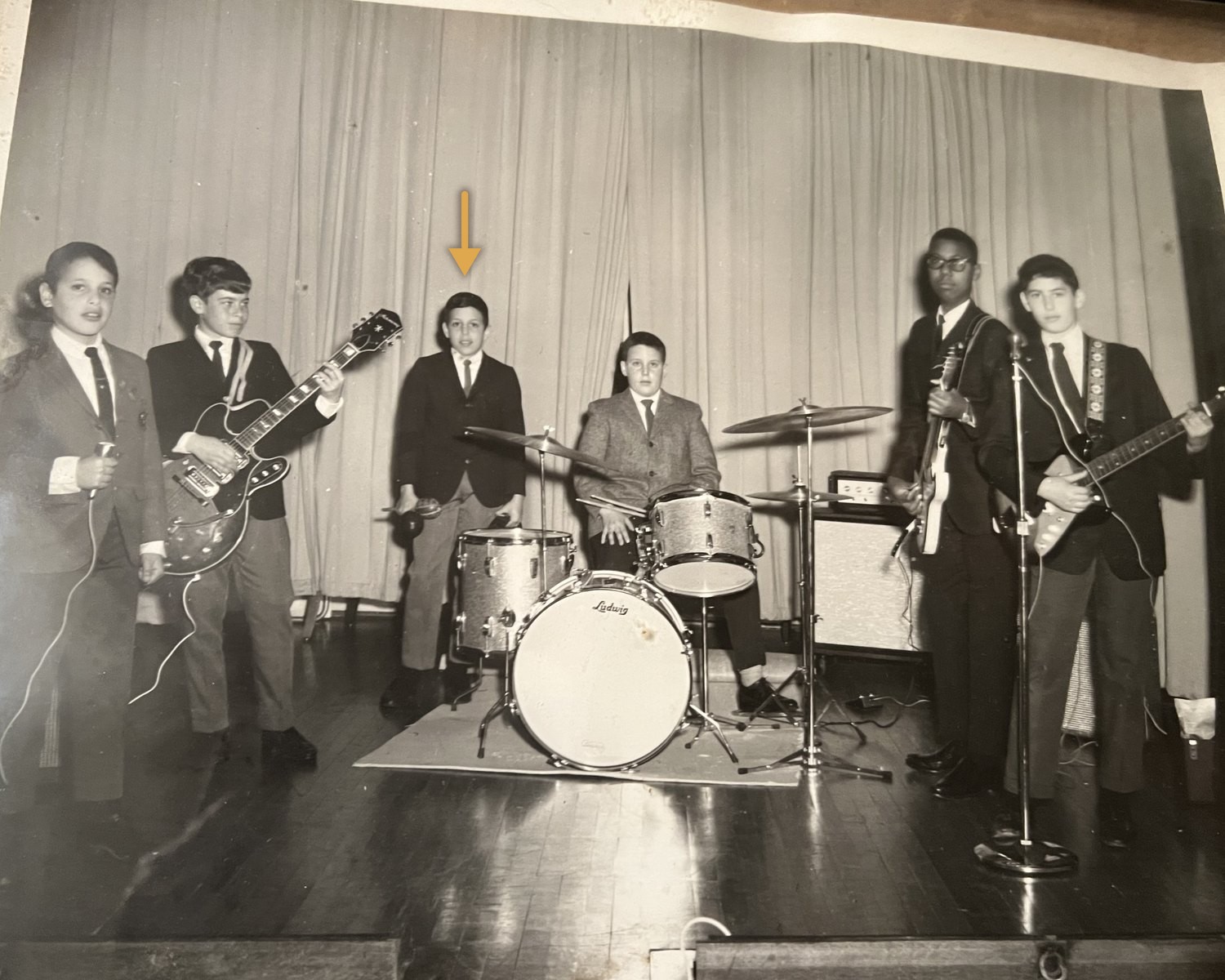 Six teenagers in jackets and ties on the Temple Emanuel stage in 1966. A small gold arrow marks David Licht, standing beside a sparkle Ludwig drum kit with a tambourine and maracas in hand.